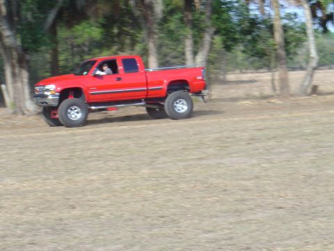 1987 Suzuki Samurai - Red lifted pickup truck with crew cab driving on a dirt field with trees in the background.
