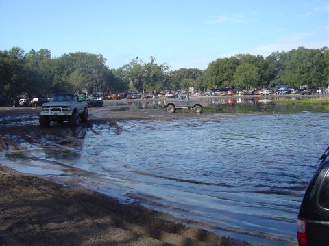 1987 Suzuki Samurai - Multiple trucks navigate a muddy and flooded terrain under a clear blue sky.