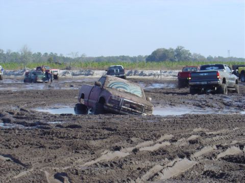 1987 Suzuki Samurai - Multiple trucks are stuck in the mud at an outdoor event, with one vehicle tilted precariously.