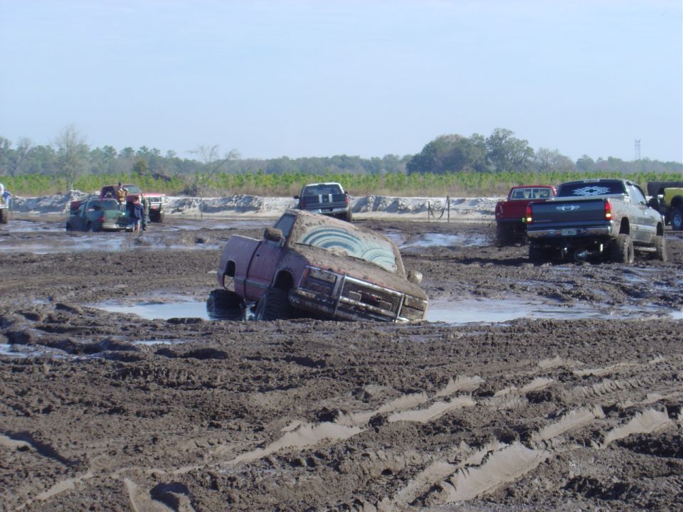 Several trucks are stuck in deep mud at an outdoor event. One truck is tilted