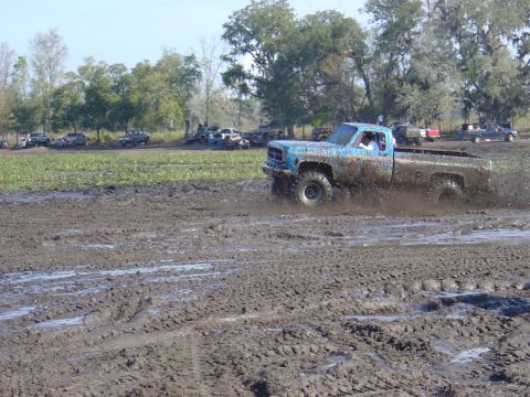 1987 Suzuki Samurai - A blue pickup truck navigates a muddy track, kicking up spray as other vehicles wait in the background.