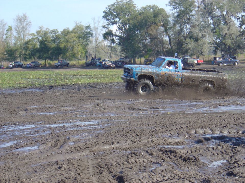 Blue pickup truck splashing through mud, other vehicles parked in background