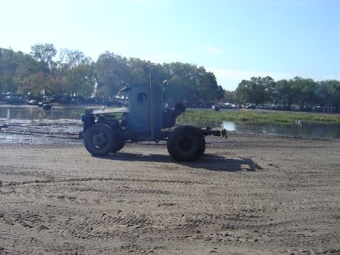 1987 Suzuki Samurai - A modified truck chassis sits on a muddy track, with trees and parked vehicles in the background.