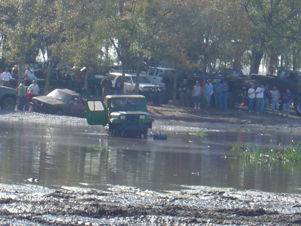Muddy scene with several off-road vehicles, one green Jeep with open door partially submerged