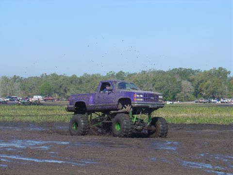 1987 Suzuki Samurai - A lifted purple pickup truck navigates a muddy track, kicking up dirt under a clear blue sky.