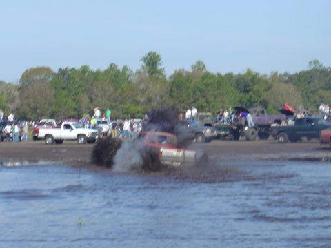 1987 Suzuki Samurai - A red truck is stuck in a mud pit during an event, with multiple other vehicles and spectators visible in the background.
