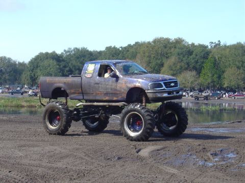 1987 Suzuki Samurai - A heavily modified pickup truck with large tires navigates a muddy terrain under a clear sky.