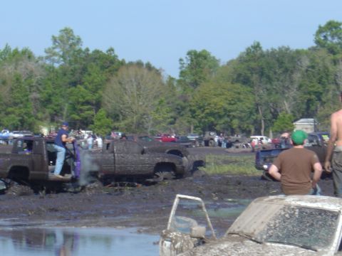 1987 Suzuki Samurai - Multiple trucks are stuck in a muddy field with spectators watching the event.
