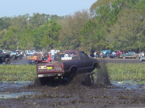 1987 Suzuki Samurai - Several vehicles are stuck in a mud pit during an event, with trees and spectators visible in the background.