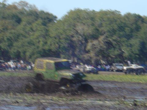 1987 Suzuki Samurai - Multiple off-road vehicles navigate a muddy track with trees and spectators visible.