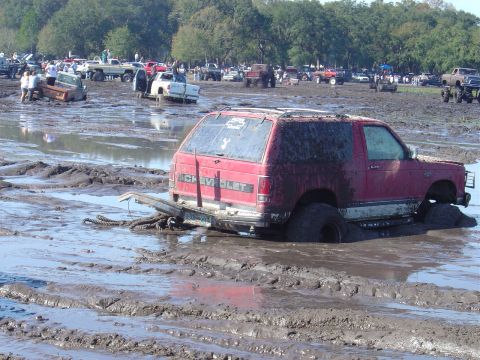 1987 Suzuki Samurai - A red Chevrolet SUV is deeply stuck in a muddy field, with other vehicles visible in the background.
