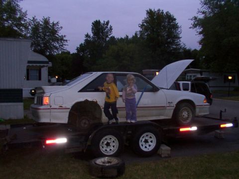 1988 Ford Mustang - Two children stand on a trailer carrying a white coupe with its hood open.