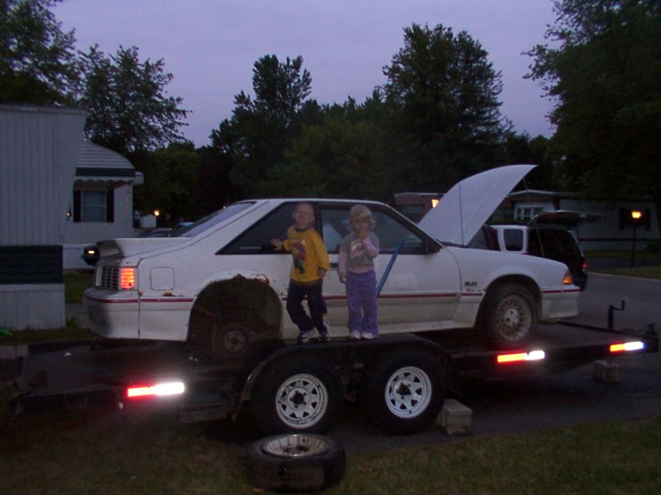 White coupe on trailer with hood up, two kids standing on trailer deck