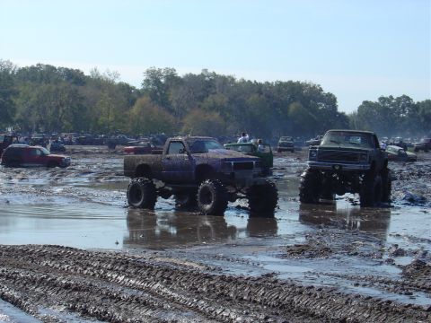 1987 Suzuki Samurai - Several modified trucks navigate a challenging mud pit under a clear sky.