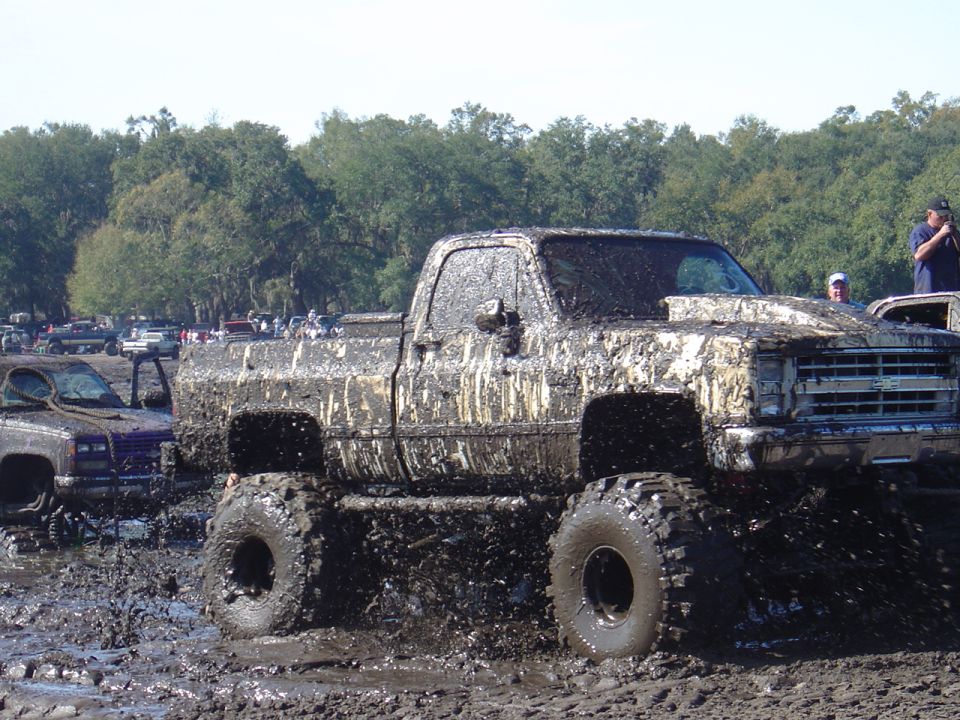 Mud-covered pickup truck with large tires in a mud pit, other vehicles in background