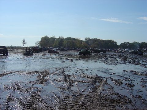 1987 Suzuki Samurai - A muddy field is filled with various vehicles navigating through the muck and water on a sunny day.