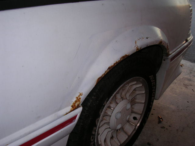 White car's rear wheel well with rust around the fender and a red stripe on the trim