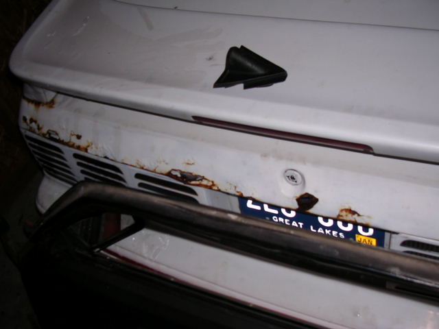 Rear of white car with rust spots, blue Great Lakes license plate, and tubular bumper bar
