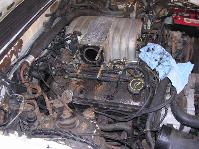 Dirty V8 engine bay with throttle body removed from intake plenum, blue rag draped at right