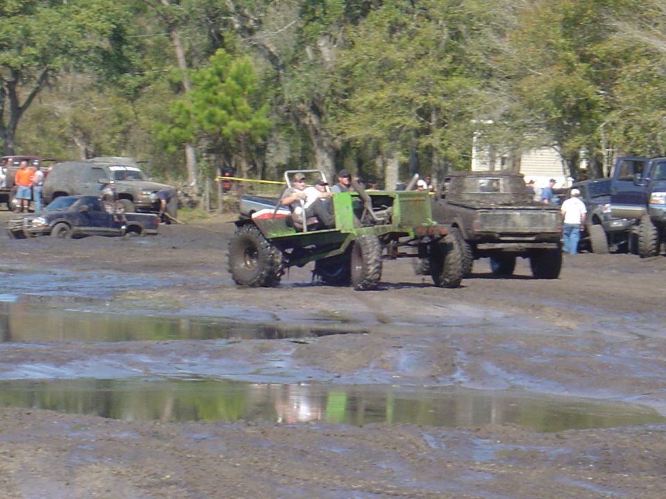 Muddy off-road vehicles with people in them driving through a muddy field with trees in the background