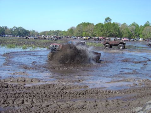 1987 Suzuki Samurai - Multiple off-road vehicles participate in a mud bogging event, with one red pickup truck prominently stuck in the mire.
