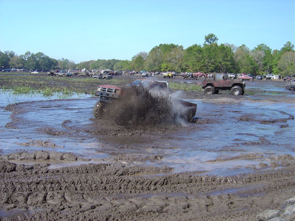 Red pickup truck spraying mud while stuck in a mud pit with other vehicles in background
