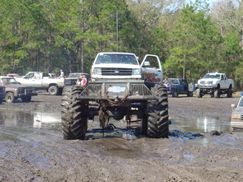 1987 Suzuki Samurai - A heavily modified monster truck sits in a muddy area with other vehicles visible in the background.