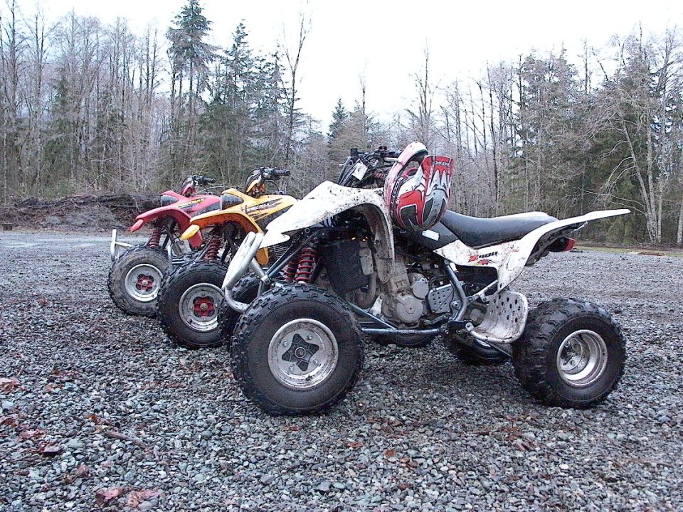 White sport ATV with helmet on seat, parked on gravel beside yellow and red ATVs among bare trees