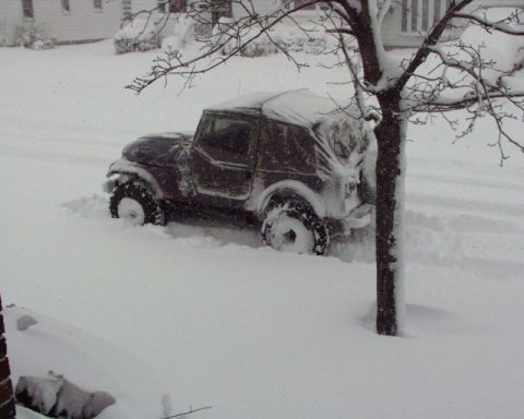 1989 Ford Bronco - A Jeep CJ-7 is stuck in deep snow on a street, with a tree in the foreground.