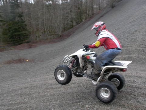 2003 Suzuki LTZ400 - A rider on a white Suzuki quad bike performs a wheelie on a loose gravel surface with trees in the background.
