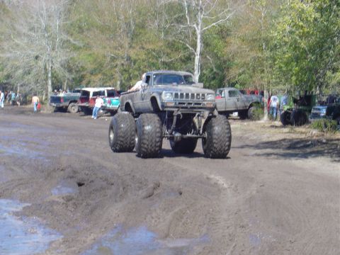 1987 Suzuki Samurai - A heavily modified truck with oversized tires navigates a muddy terrain, with other vehicles and spectators in the background.