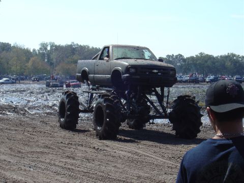 1987 Suzuki Samurai - A heavily modified pickup truck with oversized tires sits in a muddy area, with other vehicles and spectators visible in the distance.