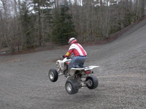 2003 Suzuki LTZ400 - A rider in a red and white jacket jumps a quad bike over a gravel surface with trees in the background.