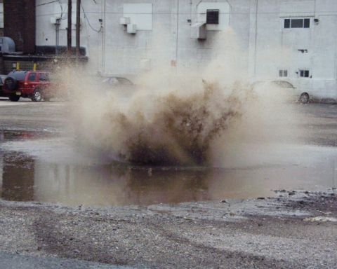 1989 Ford Bronco - A car creates a massive splash as it drives through a large puddle in a parking lot.