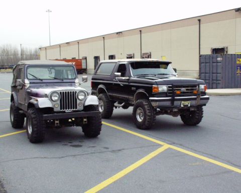 1989 Ford Bronco - A black Ford Bronco and a purple Jeep are parked side-by-side in a parking lot with a warehouse in the background.