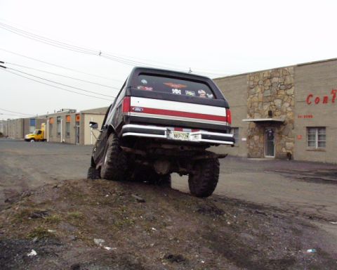 1989 Ford Bronco - 1989 Ford Bronco is shown from the rear, tilted on a dirt mound with a building in the background.