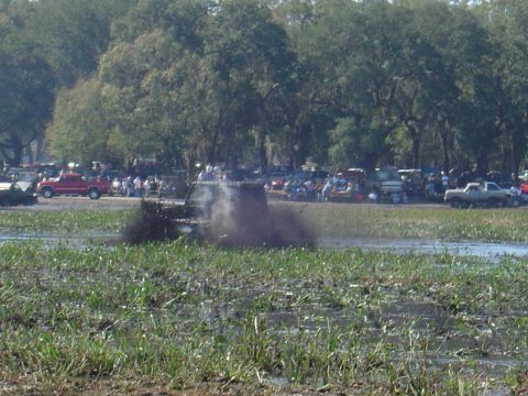1987 Suzuki Samurai - An off-road vehicle navigates through a muddy terrain, with spectators and other vehicles gathered in the background.