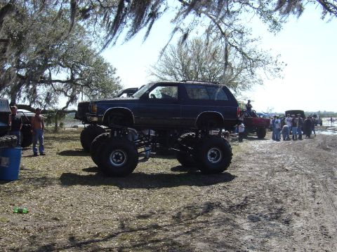 1987 Suzuki Samurai - A dark blue monster truck with oversized tires is parked on a dirt lot, with other vehicles and people in the background.