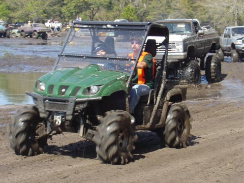 1987 Suzuki Samurai - Two people ride in a green ATV through a muddy area, followed by a lifted pickup truck.