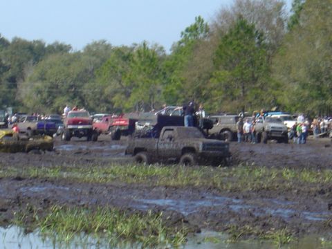 1987 Suzuki Samurai - Multiple off-road vehicles are stuck in a large mud pit during an event, with a crowd watching from the sidelines.