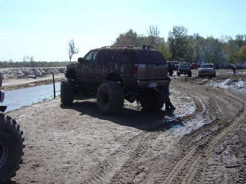 1987 Suzuki Samurai - A heavily mud-splattered SUV with massive tires navigates a muddy track, with other vehicles visible in the background.