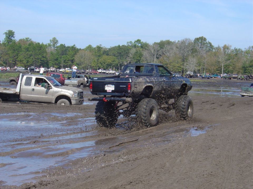 Black monster truck with large tires driving through mud, another truck stuck in the background