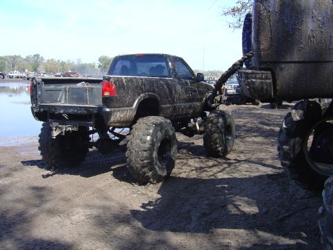 1987 Suzuki Samurai - A heavily modified black pickup truck, caked in mud, is being towed by another vehicle in a muddy outdoor area.