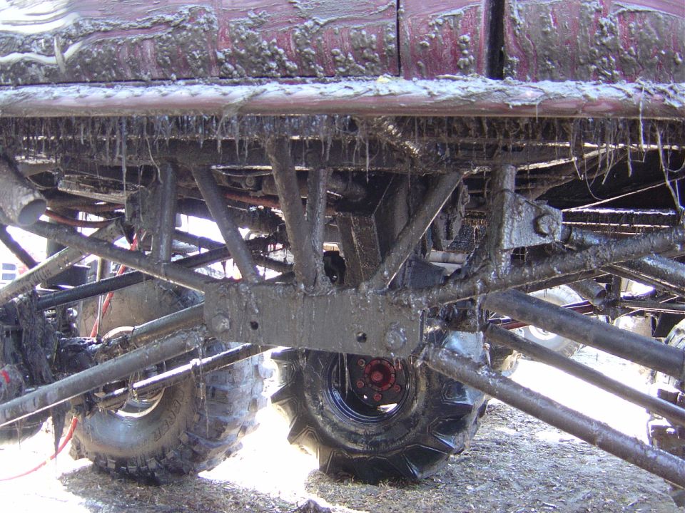 Mud-covered undercarriage of an off-road vehicle with large tires and suspension components visible. Supermotors.net logo in lower right