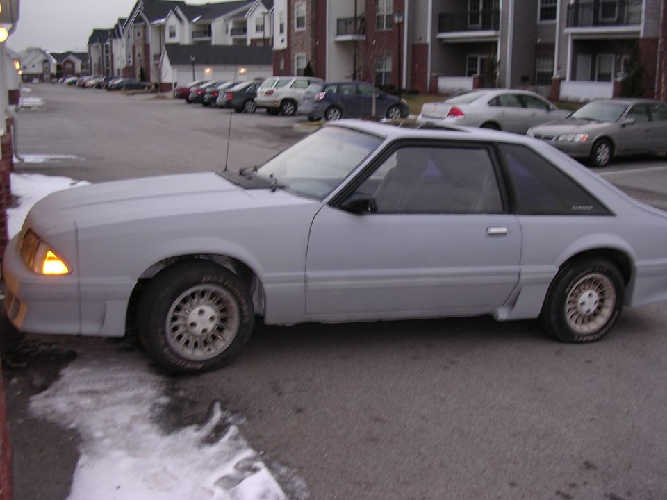 Gray Ford Mustang parked outside apartment buildings. Car is unpainted and appears to be undergoing bodywork