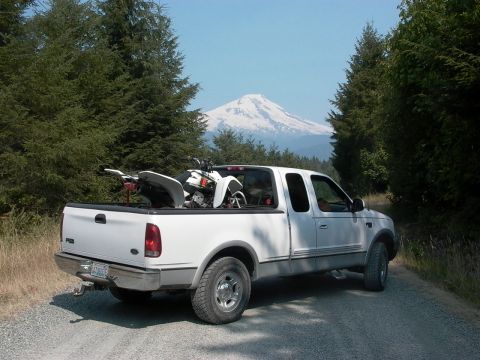 1999 Ford F-150 - A white 1999 Ford F-150 pickup truck is parked on a dirt road, carrying an ATV in its bed, with a snow-capped mountain visible in the dista…