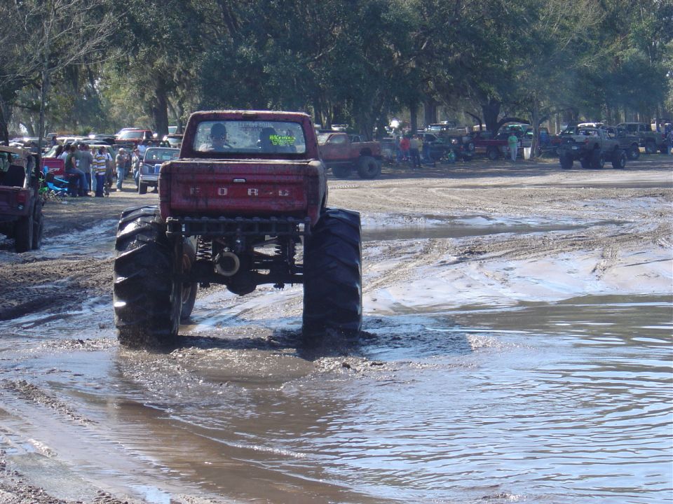 Red Ford pickup with huge tires driving through mud, other vehicles and people in background