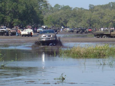 1987 Suzuki Samurai - A muddy Ford F-150 navigates through a water obstacle at an outdoor event with multiple vehicles present.