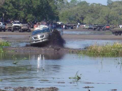 1987 Suzuki Samurai - A silver SUV navigates a muddy obstacle course, with other vehicles and spectators in the background.
