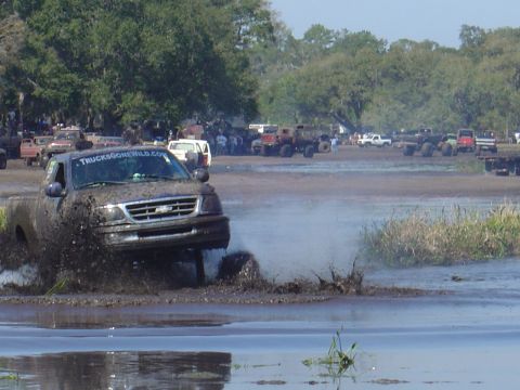 1987 Suzuki Samurai - A truck navigates a muddy track, kicking up water and mud as other vehicles and people watch from the sidelines.